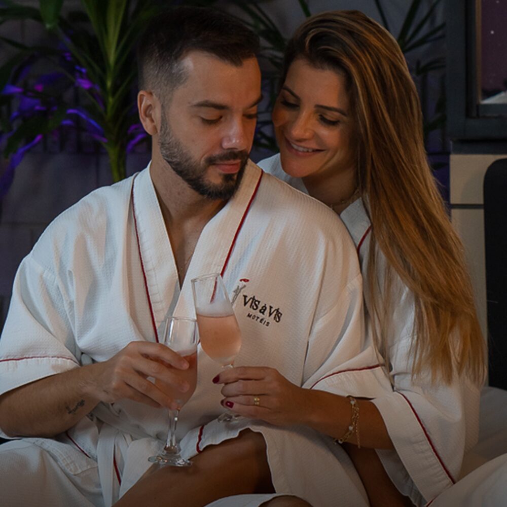 Casal brindando com taças de espumante usando os roupões do Vis à Vis para celebrar o aniversário